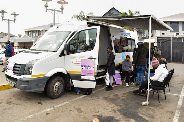 A mobile van with its side door open is parked. Several people are gathered next to it with a pop-up clinic out of the van.
