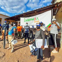A group of people stand outside wearing face masks in front of a ZAMPHIA sign under a blue sky.